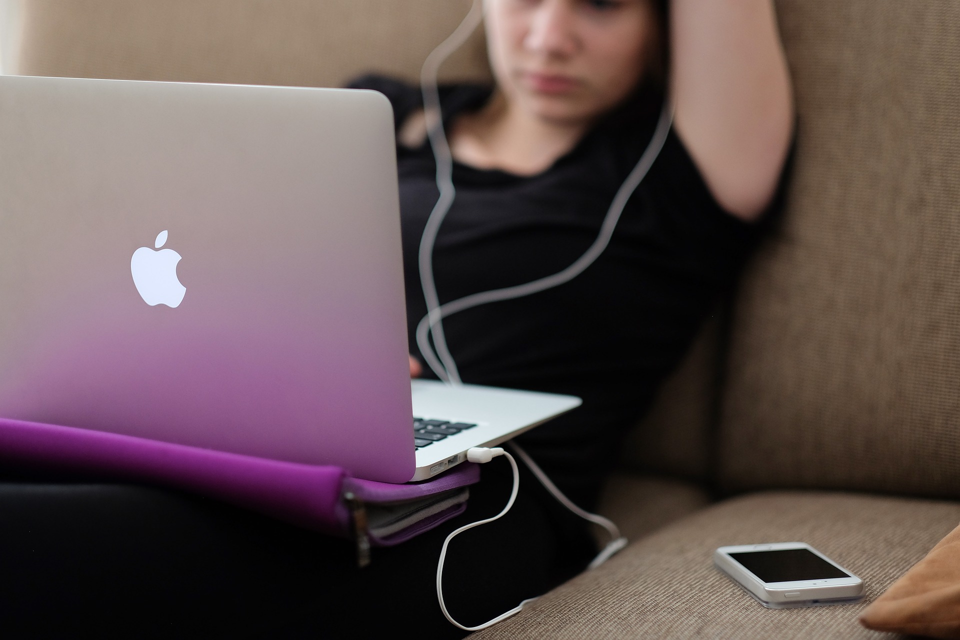 A young woman slouched on a sofa working on an Apple laptop with headphones in and a phone by her side.