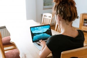 A woman with brown hair and a black shirt facing away from the camera and sitting at a dining room table working on a laptop.